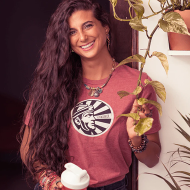 Smiling woman with long curly hair wearing a Sunwarrior shirt holding a plant and a shaker cup